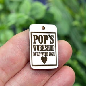 May include: A rectangular silver-coloured metal tag with a small hole at the top. The front features a dark engraved border and the text "POP'S WORKSHOP" above "BUILT WITH LOVE" and a small heart symbol centred at the bottom of the design.