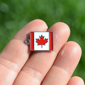 May include: A silver charm with a red maple leaf on a white background. The charm is shaped like a rectangle and has a small loop at the top for attaching to a bracelet or necklace.