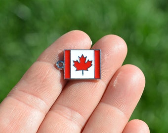 May include: A silver charm featuring a red maple leaf on a white background. The charm is rectangular in shape and has a small loop at the top for attaching to a bracelet or necklace.