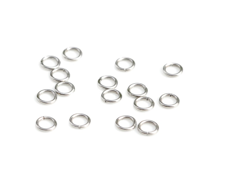 May include: A collection of silver jump rings, scattered on a white background. These rings are used for jewelry making and crafting.