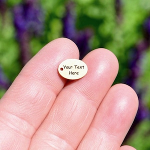 May include: A small, oval-shaped gold-colored metal charm featuring a single hole on the left side. The surface is engraved with the black text "Your Text Here" in a simple, sans-serif font, centered on the smooth metallic face.