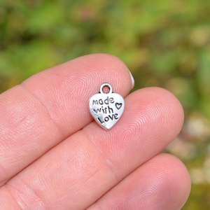 May include: A small, silver-colored heart charm with the words "made with love" engraved on the front. The charm has a small loop at the top for attaching to a necklace or bracelet. A tiny heart is next to the word "love".