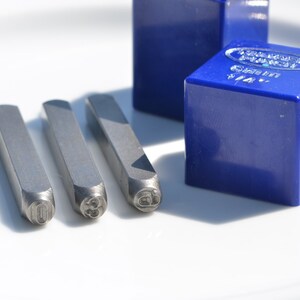May include: Four metal letter stamps with the numbers 0, 3, 6, and 9 on a white surface. The stamps are next to a blue plastic box with the text "FORGED IN ENGLAND" and "SIZE 1/8" on it.