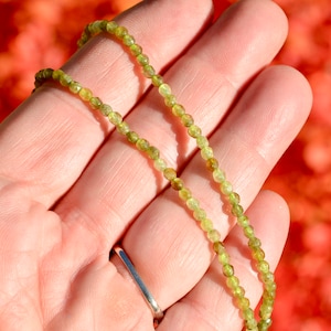 May include: A green gemstone necklace made with small, faceted beads. The necklace is displayed on a hand, with the beads forming two strands.
