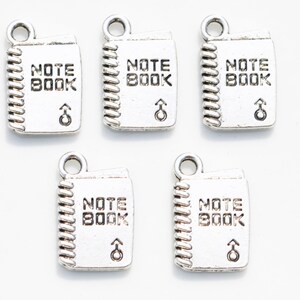 K&ouml;nnte beinhalten: F&uuml;nf silberne Anh&auml;nger in Form eines Notizbuchs mit den Worten "NOTE BOOK" und einem nach oben zeigenden Pfeil.