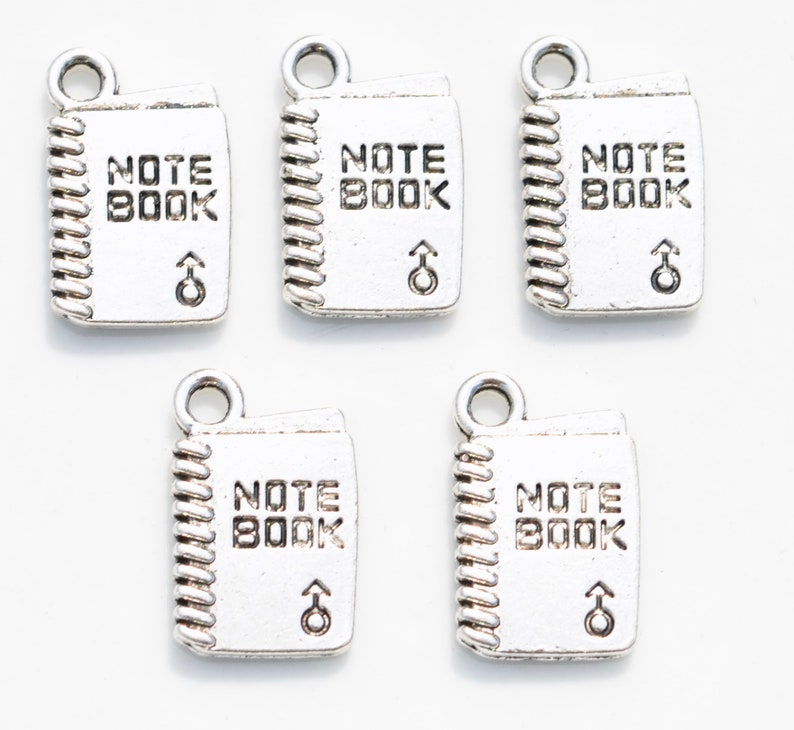 K&ouml;nnte beinhalten: F&uuml;nf silberne Anh&auml;nger in Form eines Notizbuchs mit den Worten "NOTE BOOK" und einem nach oben zeigenden Pfeil.