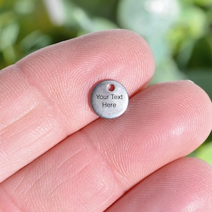 May include: A small, round, silver-coloured charm with a hole at the top. The charm has the text "Your Text Here" printed on it. The charm is held between two fingers.