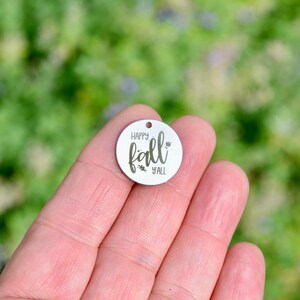May include: A small, circular, silver-toned metal charm featuring the engraved text "HAPPY fall Y'ALL" in a script font, with a small hole at the top for attachment. The charm has a smooth, reflective surface and is held against a blurred background.