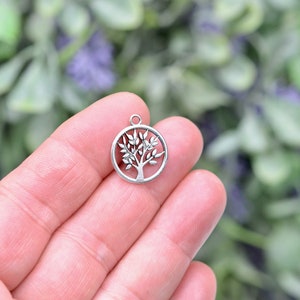 May include: Silver-toned metal charm featuring a tree of life design. The charm is round and has a hole at the top for attaching to a necklace or bracelet.