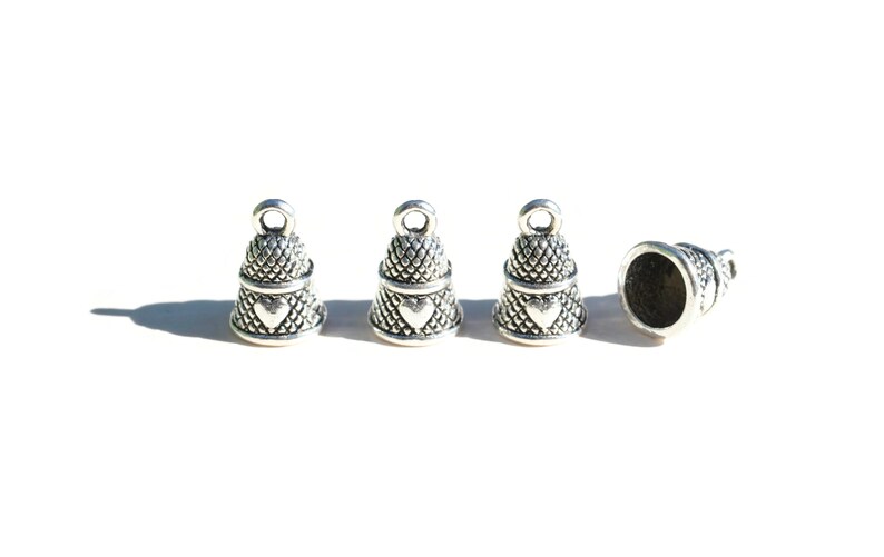 Peut inclure: Quatre breloques en forme de d&eacute; &agrave; coudre en m&eacute;tal argent&eacute; dispos&eacute;es en ligne. Chaque breloque pr&eacute;sente un motif textur&eacute; en croisillons, un petit c&oelig;ur en relief sur le c&ocirc;t&eacute; et une boucle circulaire au sommet pour la fixation.