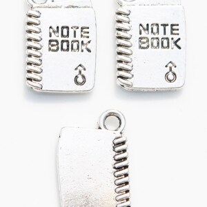 K&ouml;nnte beinhalten: Drei silberne Anh&auml;nger in Form von Notizb&uuml;chern mit Spiralbindung. Die Anh&auml;nger haben die Worte "NOTE BOOK" auf der Vorderseite eingraviert.
