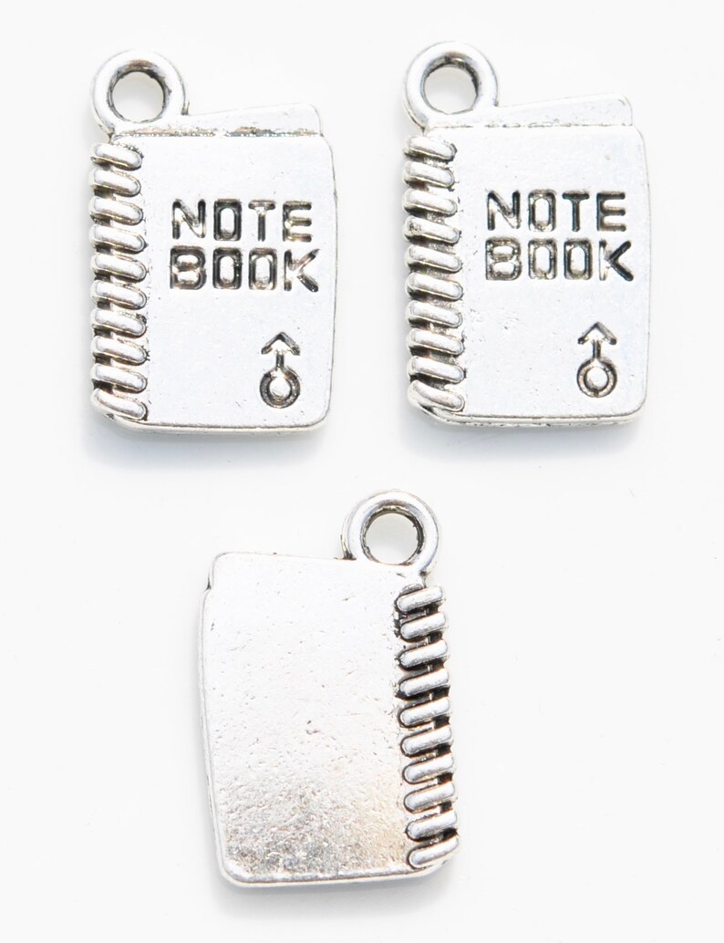 K&ouml;nnte beinhalten: Drei silberne Anh&auml;nger in Form von Notizb&uuml;chern mit Spiralbindung. Die Anh&auml;nger haben die Worte "NOTE BOOK" auf der Vorderseite eingraviert.