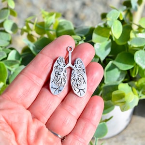 May include: Silver charm in the shape of a human lung with detailed anatomical features. The charm is held in a hand against a green leafy background.