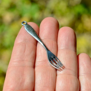 May include: A silver-toned fork charm with a loop at the top for attaching to a necklace or bracelet.