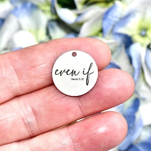 May include: A small, round, silver-colored charm with the words "even if" and "Daniel 3:18" in black script. The charm has a small hole at the top for stringing. The background is blurred with blue and white flowers.