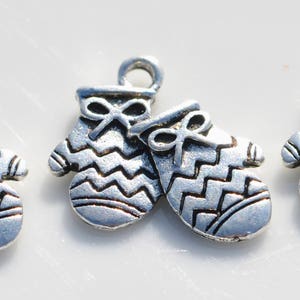 May include: Three silver-colored mitten charms with a bow detail at the top. The mittens have a zig-zag pattern and a loop for attaching to a chain or bracelet. These charms are ideal for jewelry making or crafting.