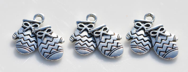 May include: Three silver-colored mitten charms with a bow detail at the top. The mittens have a zig-zag pattern and a loop for attaching to a chain or bracelet. These charms are ideal for jewelry making or crafting.
