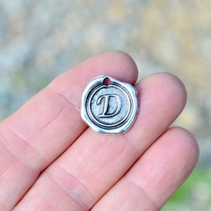 May include: Silver charm with a wax seal design featuring the letter D.