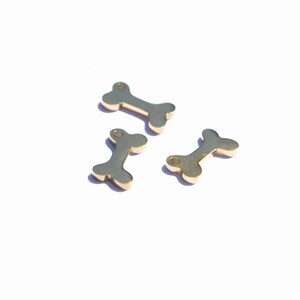 May include: Three small, gold-colored bone charms are scattered on a white surface. Each charm has a central bar with rounded ends and a hole at one end. The charms are casting shadows.