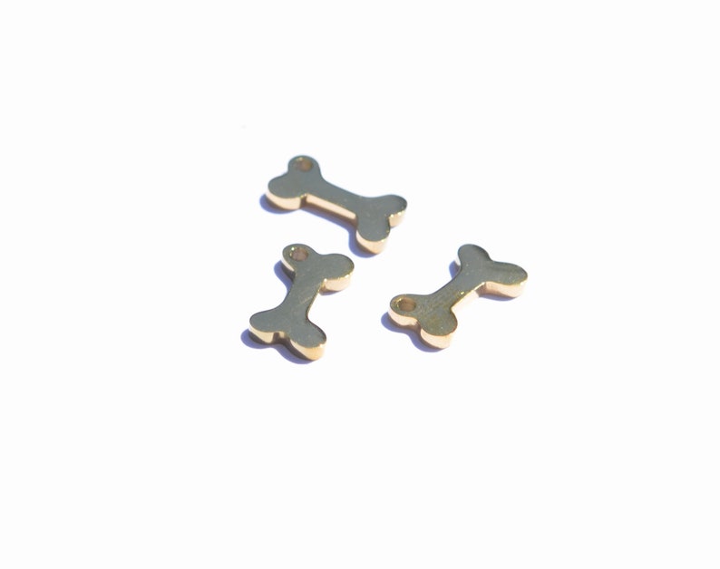May include: Three small, gold-colored bone charms are scattered on a white surface. Each charm has a central bar with rounded ends and a hole at one end. The charms are casting shadows.