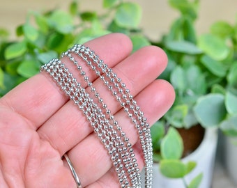 May include: Close-up of several strands of silver ball chain. The chains are held in a hand, with a blurred background of green plants. The chains are made of small, evenly spaced, metallic spheres.