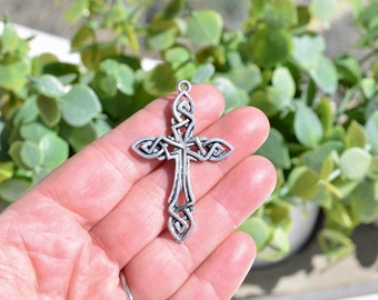 May include: A silver Celtic knot cross pendant with intricate details. The cross is held in a hand against a green leafy background.
