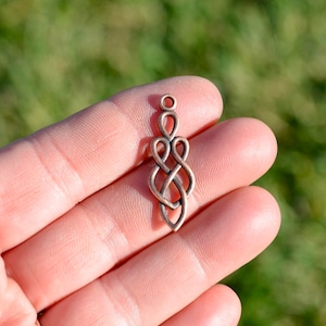 May include: A copper-colored pendant with a Celtic knot design. The pendant is elongated with a loop at the top and a pointed bottom. The intricate knotwork features interwoven loops and curves, creating a symmetrical pattern. The pendant is held in a hand against a blurred green background.