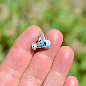 May include: Silver charm in the shape of a fish. The fish has a detailed design and a loop at the top for attaching to a necklace or bracelet.