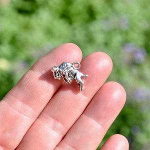 May include: Silver charm in the shape of a buffalo, the buffalo is standing on its hind legs with its head turned to the side.