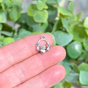 May include: Silver charm in the shape of a Claddagh ring with two hands holding a heart. The charm has a loop at the top for attaching to a necklace or bracelet.