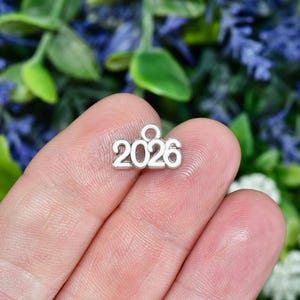 May include: A small, silver charm with the number "2026" displayed prominently. The charm is held between fingers against a backdrop of green and purple foliage. The charm is likely used for jewelry making or as a decorative element.