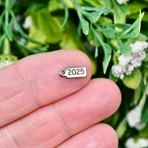 May include: A small, rectangular silver charm with the number "2025" printed in black. The charm has a small hole at one end. The charm is held in a person's hand, with a background of green leaves and white flowers.