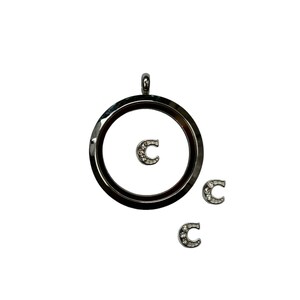 May include: A silver locket with a clear glass insert and a small silver charm with a letter C and rhinestones. Three additional charms with the letter C and rhinestones are scattered around the locket.