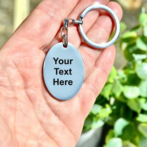 Puede incluir: Un llavero ovalado azul claro con el texto "Your Text Here" en negro. El llavero tiene un anillo y una cadena de color plateado. El fondo est&aacute; borroso, con follaje verde visible.