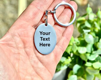 May include: A light blue oval keychain with the text "Your Text Here" in black. The keychain has a silver-colored ring and chain. The background is blurred, with green foliage visible.