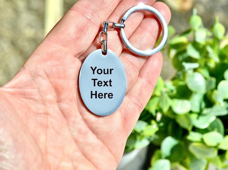 Puede incluir: Un llavero ovalado azul claro con el texto "Your Text Here" en negro. El llavero tiene un anillo y una cadena de color plateado. El fondo est&aacute; borroso, con follaje verde visible.