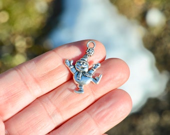 May include: Silver charm of a snowman wearing a hat and scarf, with a smiling face, holding a stick in one hand and a ball in the other. The charm is on a finger.