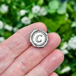 May include: A silver-colored charm with the letter "C" in the center. The charm has a circular shape with a raised border, resembling a wax seal. It has a small hole at the top for attaching to a chain or other jewelry component.