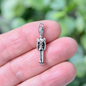 May include: A miniature silver-coloured charm depicting a toy soldier. The soldier is wearing a detailed uniform with crossed straps and a tall hat. The charm has a small loop at the top for attaching to jewellery. The background is blurred.