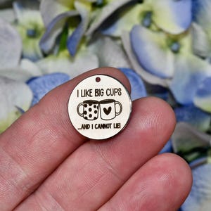 May include: A small, round wooden charm with the phrase "I LIKE BIG CUPS...AND I CANNOT LIE!" etched on it. The charm features two coffee mugs, one with polka dots and the other with a heart. A small hole is at the top for hanging.