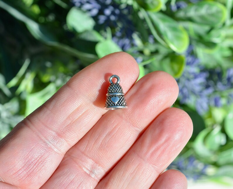 Peut inclure: Une petite breloque en m&eacute;tal argent&eacute; en forme de d&eacute; &agrave; coudre, pr&eacute;sentant un motif textur&eacute; en croisillons sur toute sa surface et un d&eacute;tail lisse en forme de c&oelig;ur centr&eacute; sur le devant. La breloque poss&egrave;de une petite boucle circulaire au sommet.