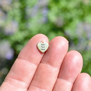 May include: A small, heart-shaped charm with a loop at the top. The charm is a light gold color and features the text "Your Text Here" in black. The charm is held between two fingers against a blurred green and purple background.