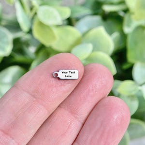 May include: A small, rectangular silver charm with a loop at one end. The charm has the text "Your Text Here" printed on it. The background is a blurred image of green leaves.