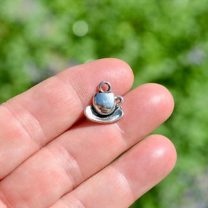 May include: A small, silver-colored charm in the shape of a teacup and saucer. The charm has a loop at the top for attaching to a bracelet or necklace. The charm is made of metal and has a shiny finish. The charm is a miniature representation of a teacup and saucer.