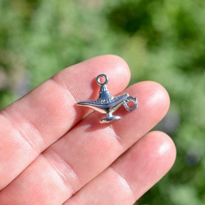 May include: A silver-toned metal charm shaped like an Aladdin's lamp. The charm has a loop at the top for attaching to a necklace or bracelet.