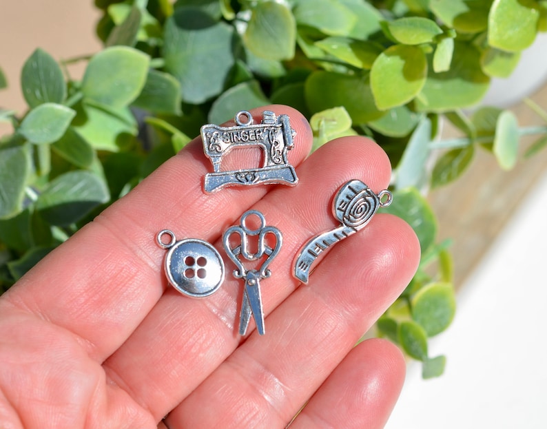 May include: A set of silver-toned metal charms featuring a sewing machine, a button, a pair of scissors, and a measuring tape. The charms are ideal for adding a touch of vintage charm to jewellery or crafts.