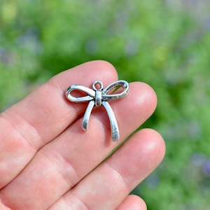 May include: Silver charm in the shape of a bow. The bow is tied with a knot in the centre and has a loop at the top for attaching to a necklace or bracelet.