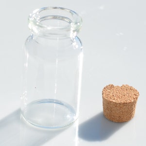 May include: A clear glass bottle with a cylindrical body and a rounded lip, next to a cork stopper. The bottle is empty and sits on a white surface. The cork is a light brown color and has a textured surface.