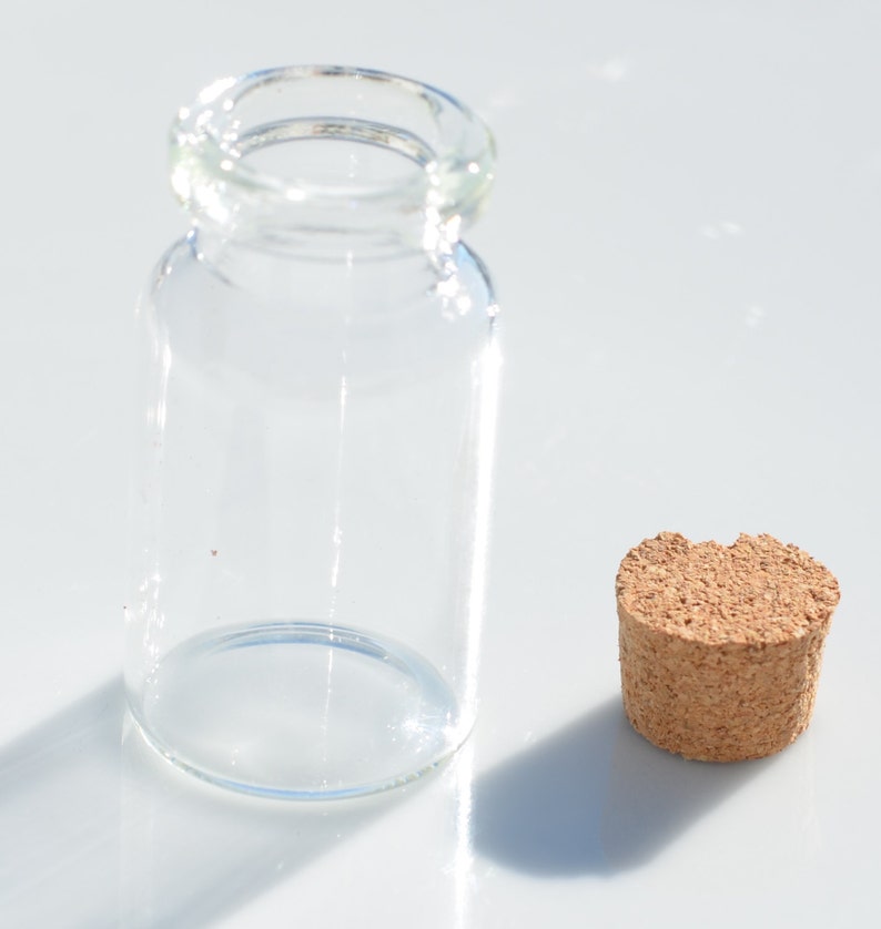 May include: A clear glass bottle with a cylindrical body and a rounded lip, next to a cork stopper. The bottle is empty and sits on a white surface. The cork is a light brown color and has a textured surface.