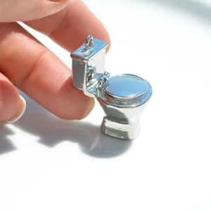 May include: A silver-colored miniature toilet charm. The toilet is open and the lid is up. The charm is small enough to fit on a finger.